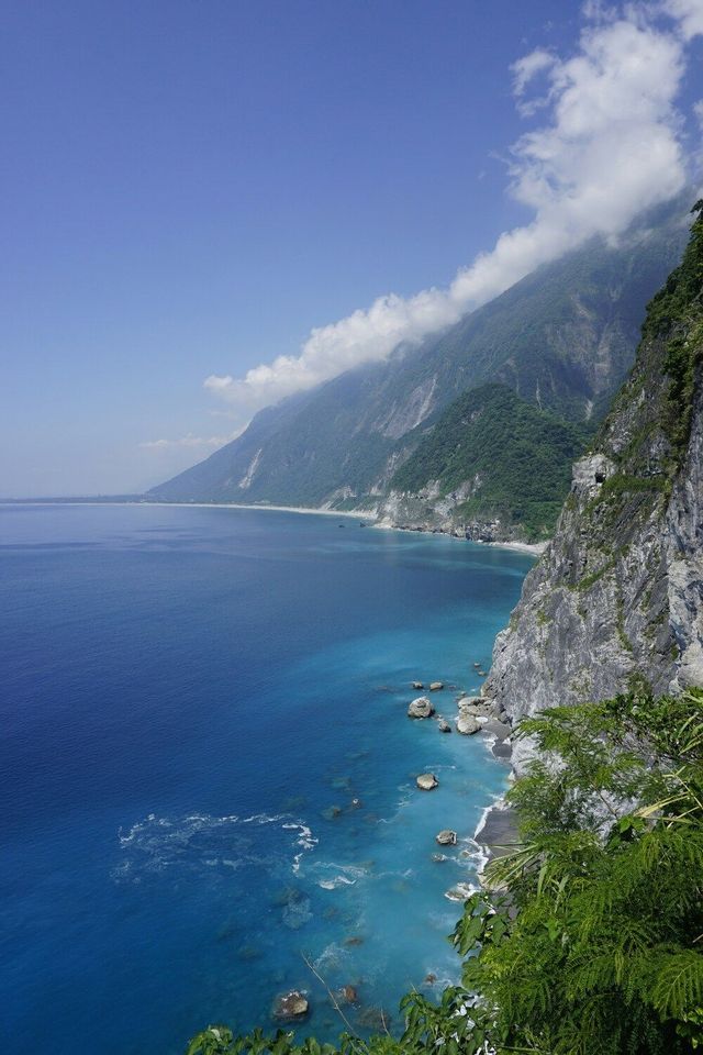 Una costa frastagliata dove ripide montagne verdi incontrano il mare vibrante blu e turchese sotto un cielo parzialmente nuvoloso.