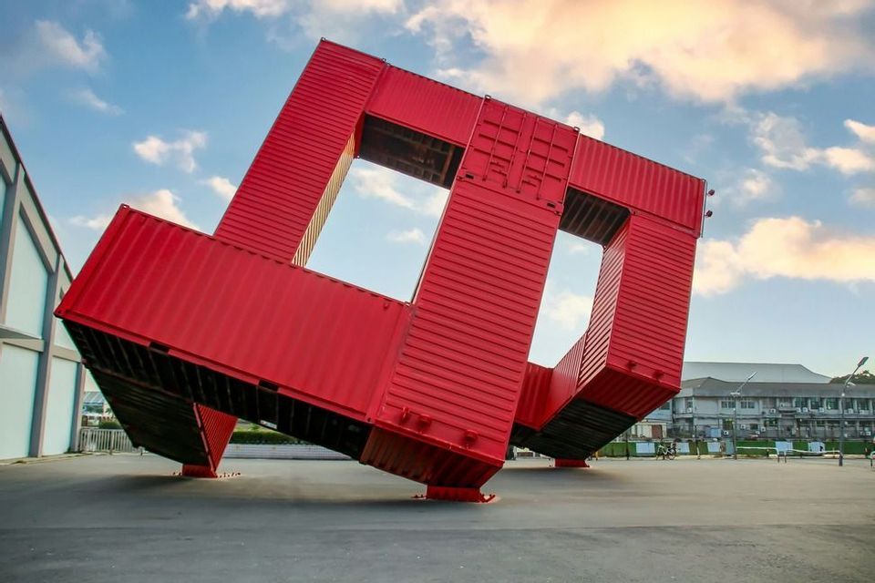 Una grande scultura rossa, fatta di container, è in equilibrio obliquo in una piazza pavimentata sotto un cielo blu con nuvole.