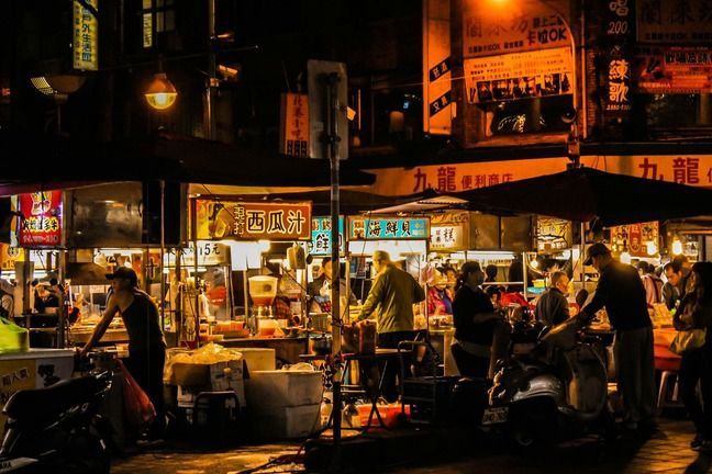Un vivace mercato notturno asiatico, con persone radunate attorno a bancarelle di cibo illuminate sotto insegne luminose.