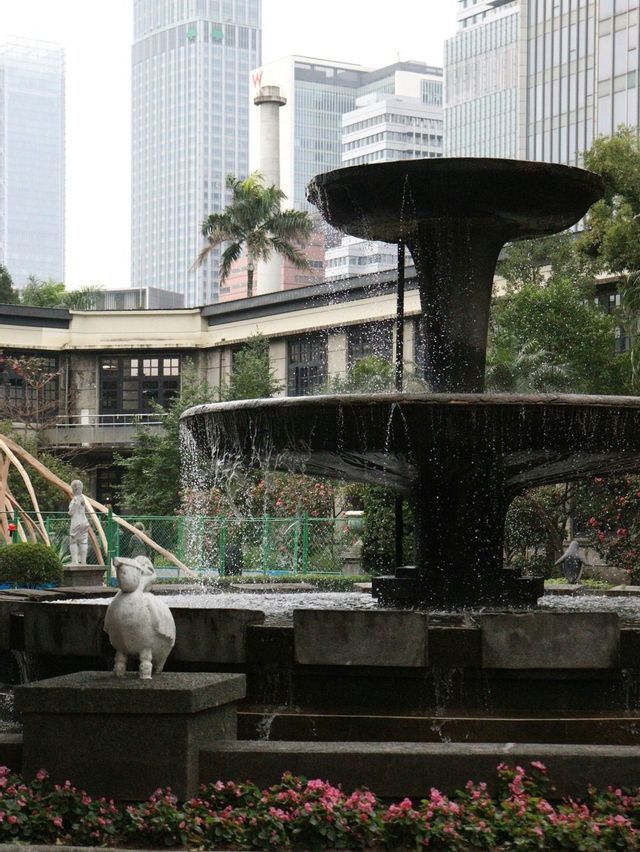 Una fontana scura a più livelli e una piccola statua di anatra bianca in un parco cittadino con moderni grattacieli sullo sfondo.
