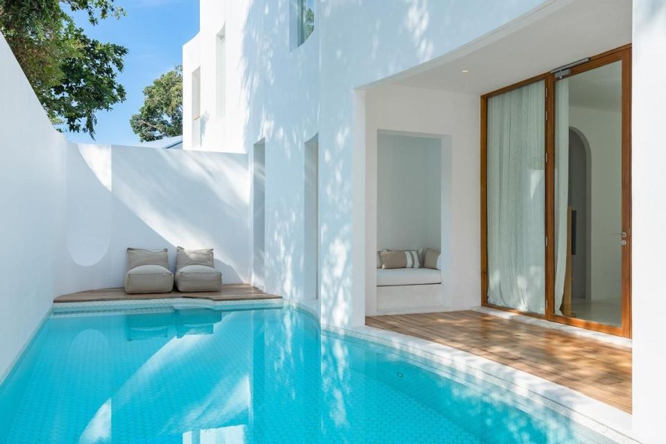 A sunlit private pool and wooden deck adjacent to a modern white building with minimalist seating.