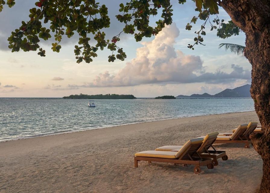 Empty sun loungers rest on a sandy beach facing the sea at dusk, with a boat and islands visible in the distance under a cloudy sky.