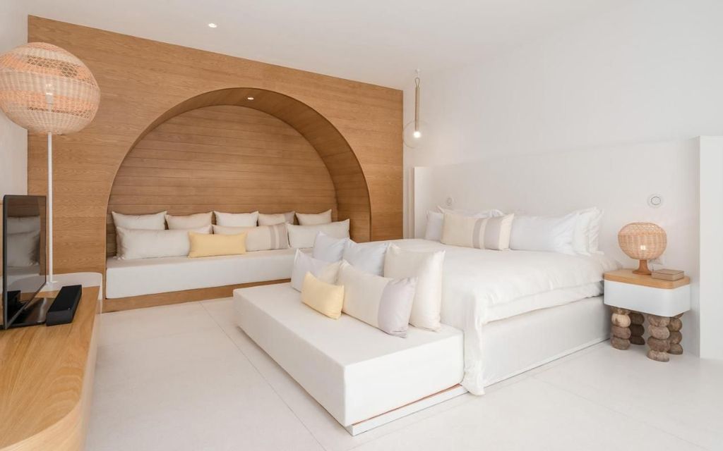 A minimalist white bedroom with a large bed and a wooden arched wall with a built-in sofa, decorated with light-colored pillows.