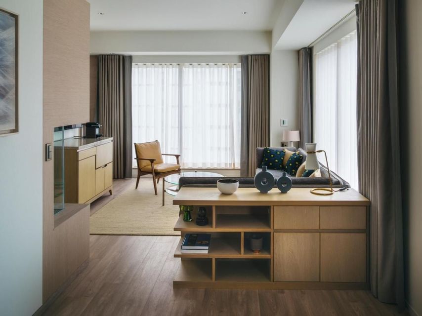 A modern living room featuring light wood furniture, a grey sofa, and large windows with sheer and heavy drapes.