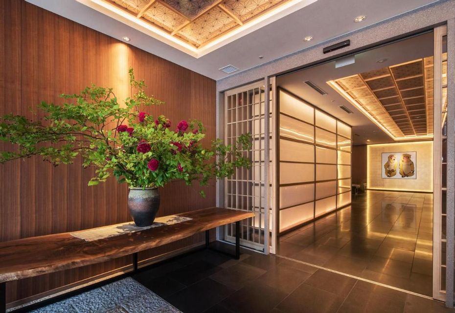 A large floral arrangement in a ceramic vase sits on a wooden bench in a Japanese-style lobby next to a warmly lit hallway.