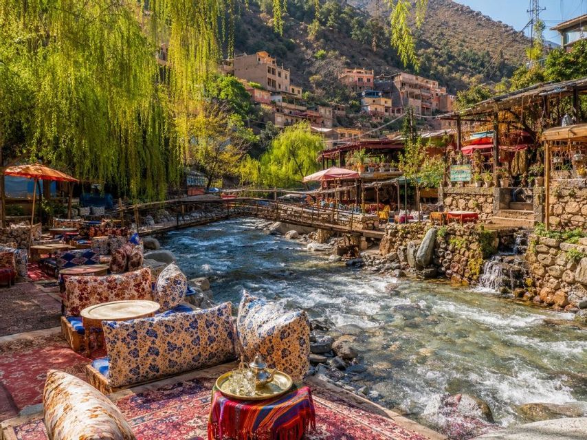 Un pintoresco café al aire libre con cojines y alfombras de colores se encuentra junto a un río caudaloso, con un pueblo en la ladera de fondo.