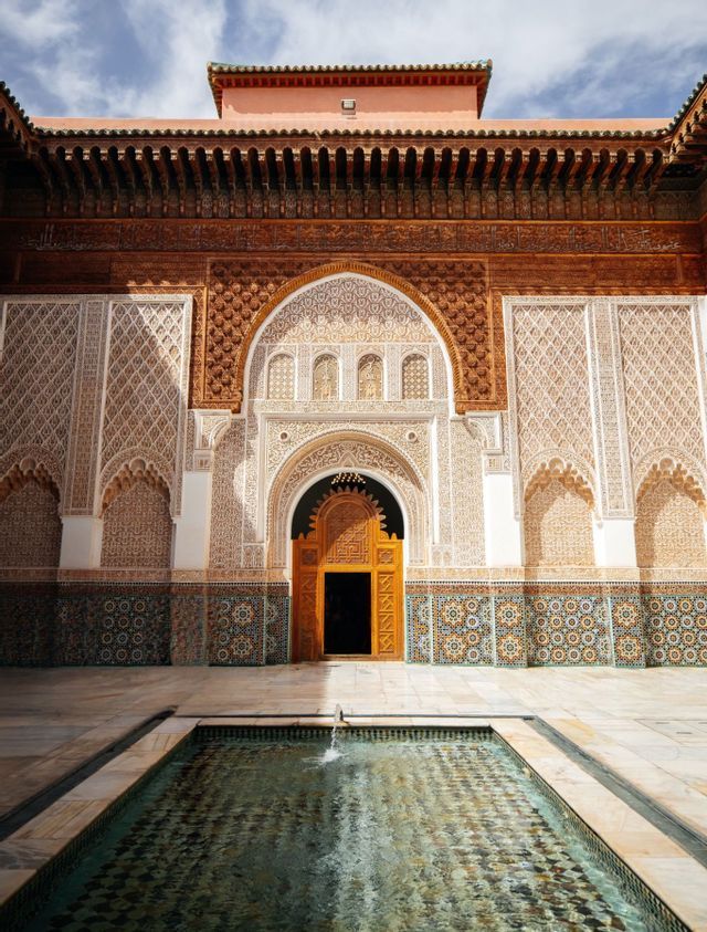 Un patio de piedra con una piscina central frente a un edificio ornamentado con puertas de madera talladas y azulejos intrincados.