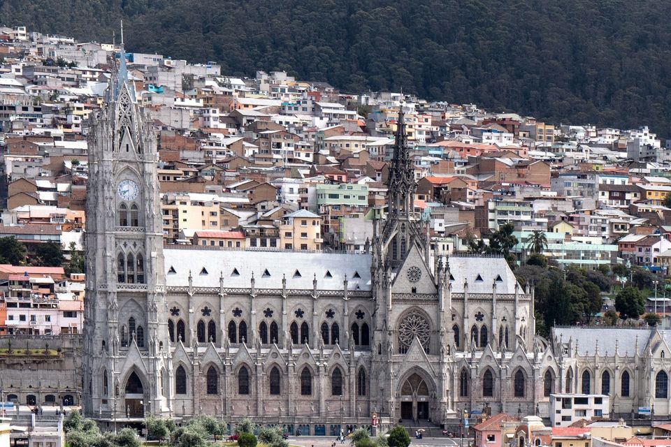 Una gran catedral gótica gris con una torre del reloj se alza frente a una densa ciudad construida en una colina boscosa.