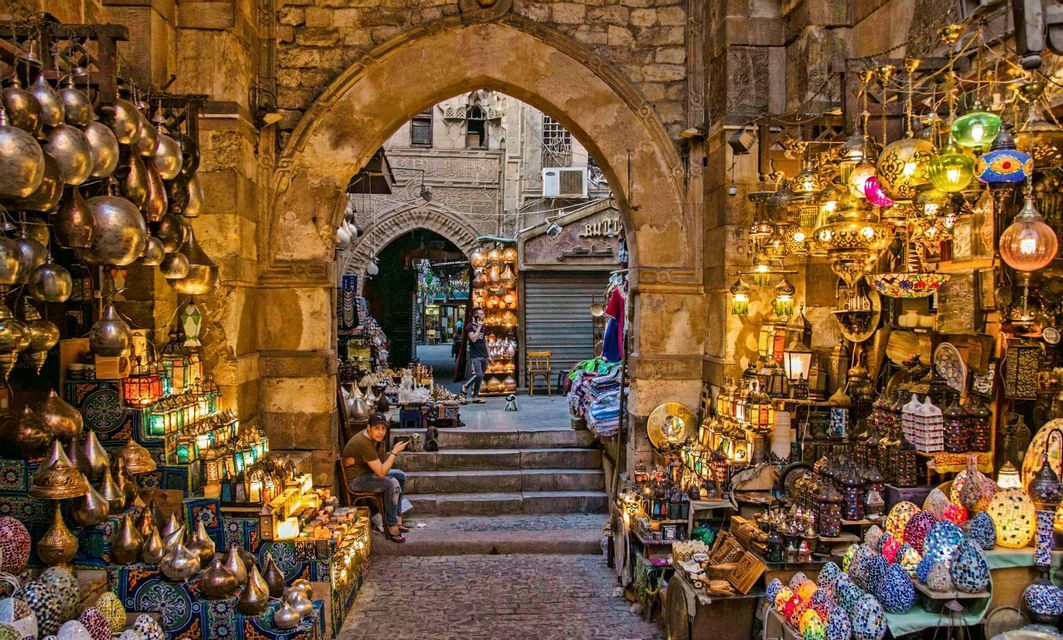 Une allée en pierre dans un marché animé est remplie d'étals présentant de nombreuses lanternes colorées en verre et en métal illuminées.