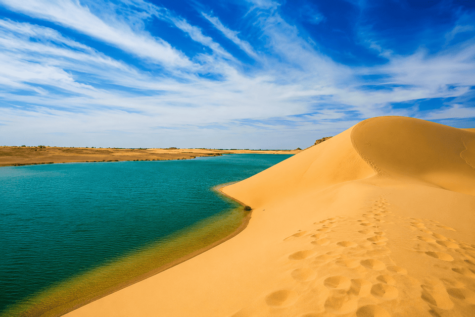 Une grande dune de sable doré avec des empreintes de pas rencontre un plan d'eau turquoise dans le désert sous un ciel bleu avec des nuages.
