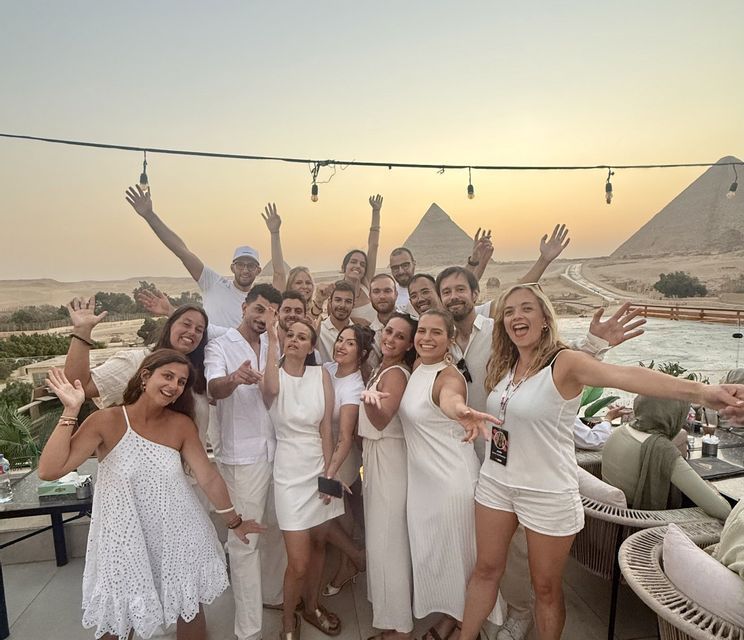 Un groupe WeRoad, habillé de blanc, pose sur une terrasse avec les pyramides de Gizeh en arrière-plan, au coucher du soleil.