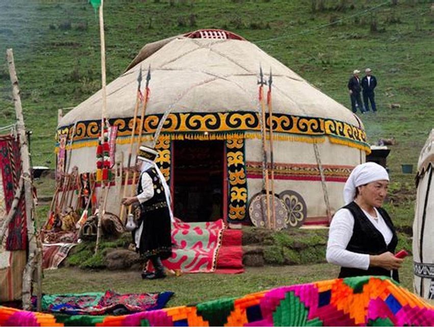 Menschen in traditioneller Kleidung stehen vor einer verzierten Jurte auf einem grasbewachsenen Feld, umgeben von Speeren und farbenfrohen Textilien.