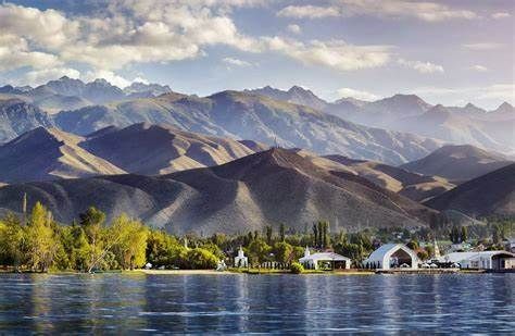 Un lago en calma se extiende frente a una orilla con árboles verdes y edificios blancos, con una vasta y ondulante cordillera de fondo bajo un cielo parcialmente nublado.