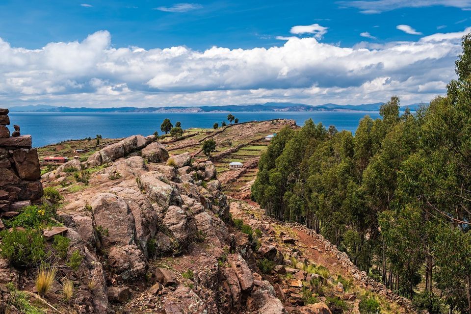 Una ladera rocosa con campos verdes aterrazados y árboles domina un gran lago azul bajo un cielo parcialmente nublado.