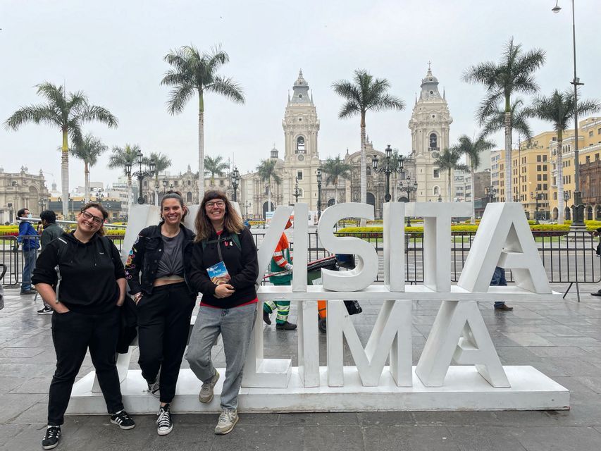 Tre donne in viaggio di gruppo WeRoad sorridono e posano accanto a un grande cartello bianco "Visita Lima" in una piazza cittadina con una cattedrale sullo sfondo.