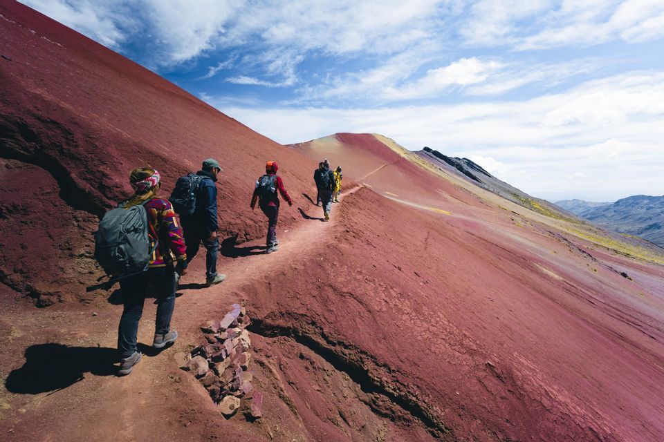Un viaggio di gruppo WeRoad facendo trekking su uno stretto sentiero lungo il fianco di una vasta montagna rossa sotto un cielo blu parzialmente nuvoloso.
