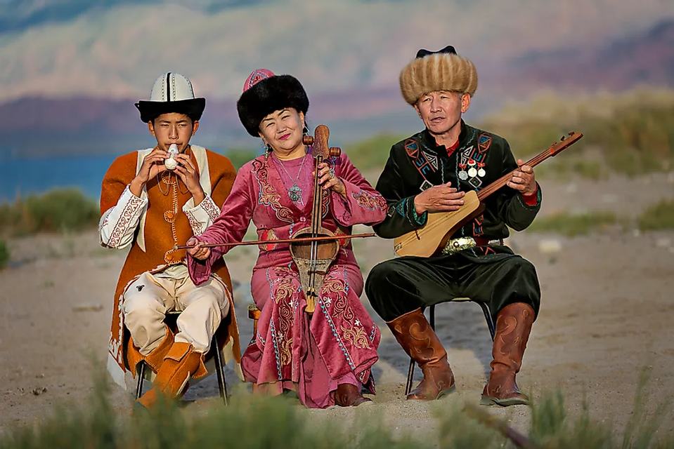 Drei Musiker in farbenfroher Tracht sitzen im Freien und spielen Saiten- und Blasinstrumente vor einer Bergkulisse.