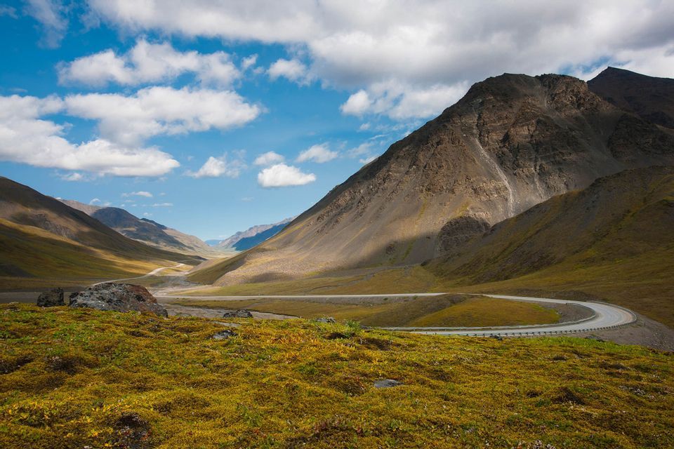 Una strada tortuosa attraversa una vasta valle di montagna coperta di tundra verde e gialla sotto un cielo blu con nuvole bianche.