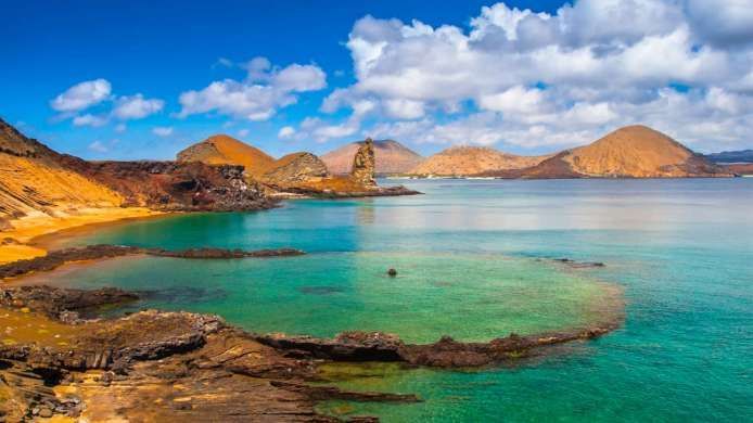 Una costa volcánica con una playa de arena que se encuentra con aguas turquesas y cristalinas, con colinas distantes bajo un cielo azul parcialmente nublado.