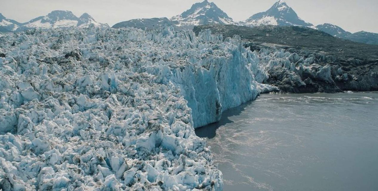 L'estremità di un enorme ghiacciaio dalla superficie irregolare incontra uno specchio d'acqua di fronte a una catena di montagne innevate.