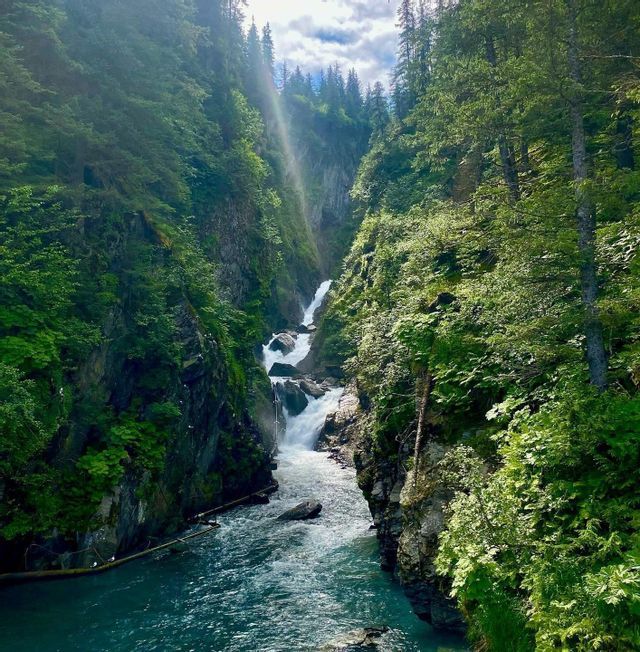 Una cascata a più livelli si getta da una gola ripida e boscosa in un fiume turchese e vibrante sottostante, con un raggio di sole che filtra tra gli alberi.