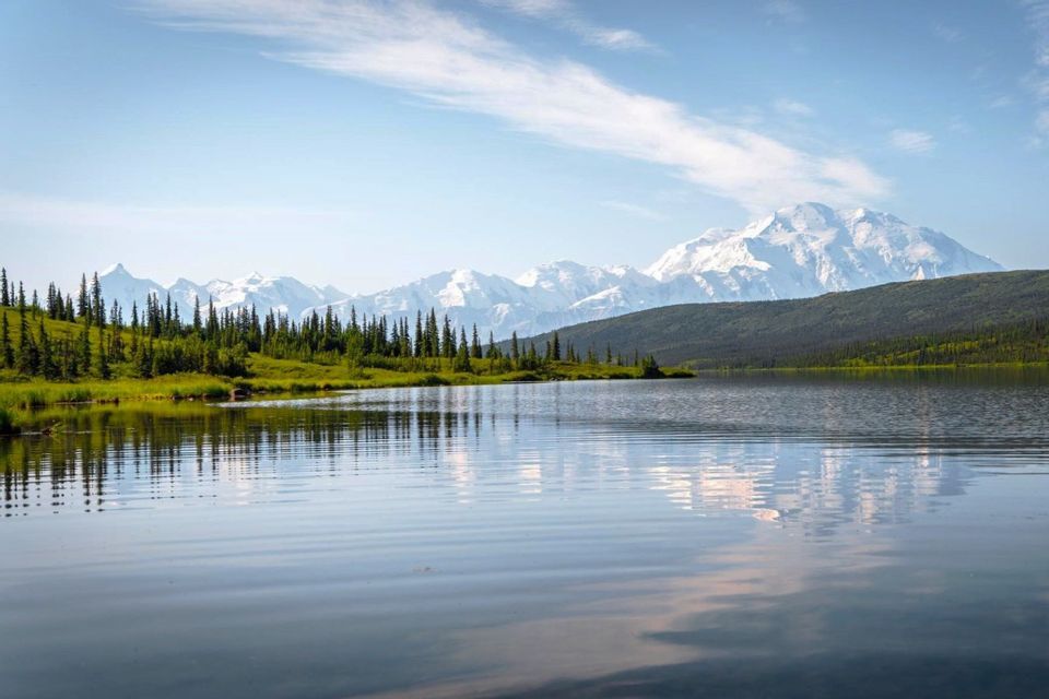 Un lago sereno riflette una fila di alberi sempreverdi e una catena montuosa innevata in lontananza sotto un cielo azzurro.