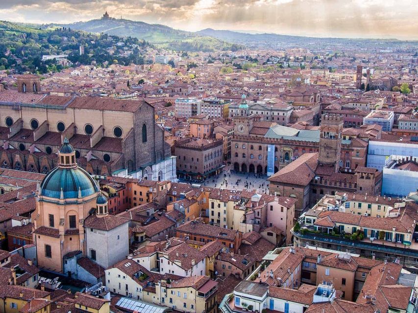 Vista aerea di una città storica con tetti di tegole rosse, una grande basilica, una cupola verde e una piazza pubblica, con colline sullo sfondo.