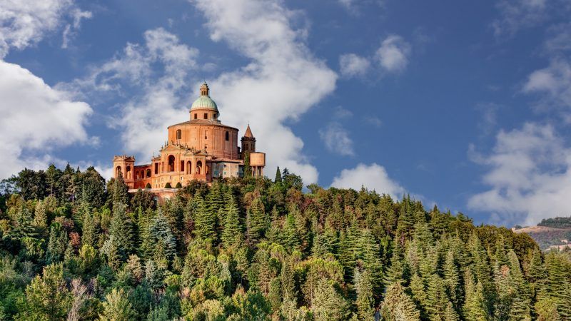 Una grande basilica color arancio con una cupola verde si erge su una collina ricoperta da fitti alberi verdi sotto un cielo parzialmente nuvoloso.