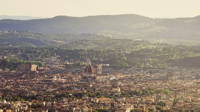 Una vista aerea di una città storica con una grande cupola di cattedrale al suo centro, sullo sfondo di dolci colline verdi.