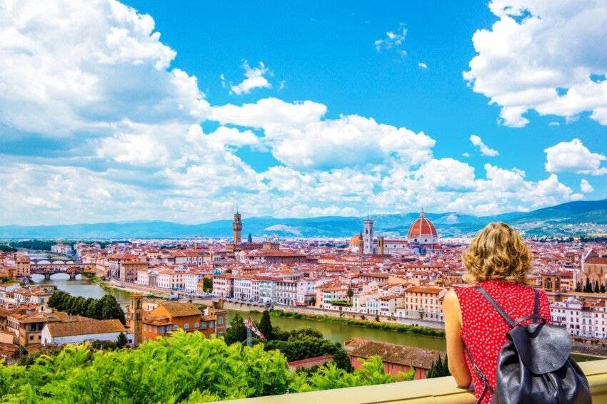 Una donna con uno zaino osserva da un punto panoramico una città storica con una grande cupola e un fiume sotto un cielo blu nuvoloso.