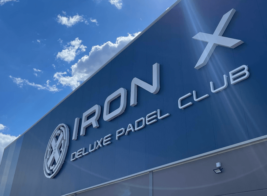 Inquadratura dal basso dell'insegna 'Iron X Deluxe Padel Club' con scritte bianche su un edificio blu, sotto un cielo parzialmente nuvoloso.