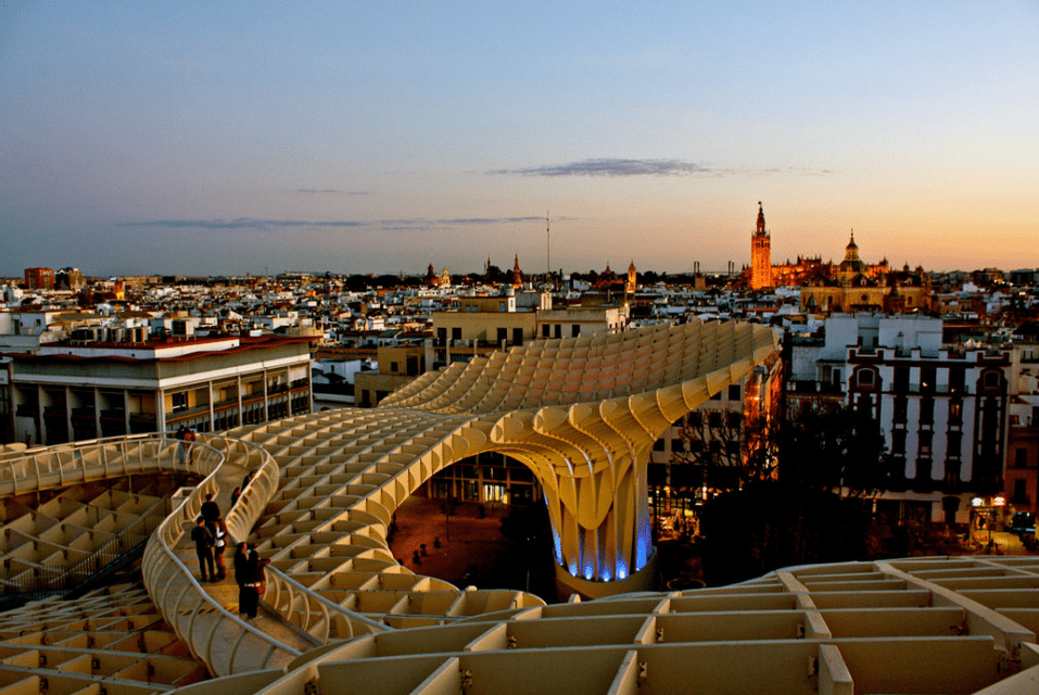 Una vista dal tortuoso camminamento di una grande e moderna struttura in legno che si affaccia su un paesaggio urbano con una cattedrale illuminata al tramonto.