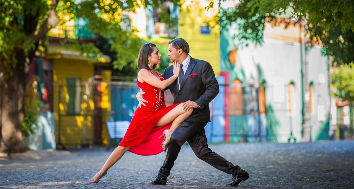 Un hombre y una mujer realizan una pose de tango en una calle empedrada, con edificios coloridos de fondo.
