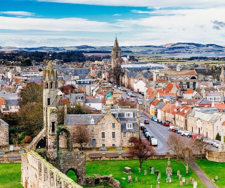 Vue en plongée d'une ville historique, avec des ruines d'une église en pierre et un cimetière au premier plan, s'étendant vers des collines lointaines.