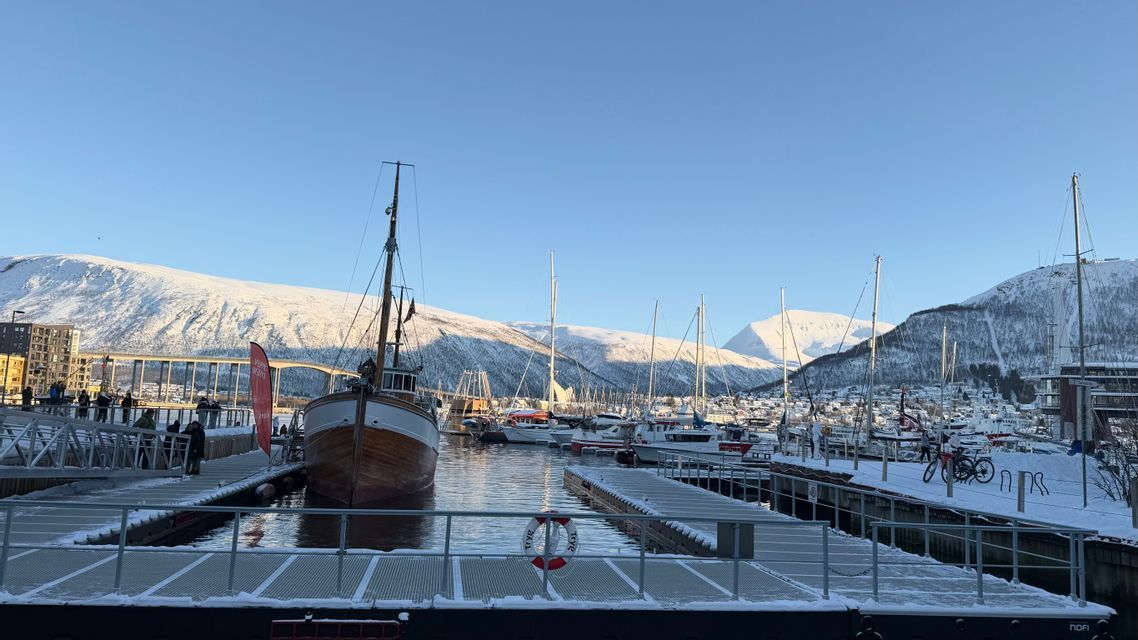 A traditional wooden boat is docked in a snowy harbor with mountains in the background under a clear, sunny sky.