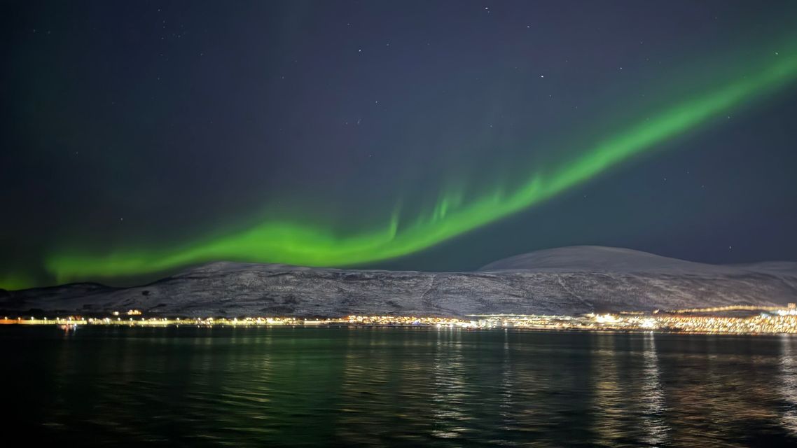 A green aurora borealis glows in the starry night sky above snowy mountains and a town reflecting on the water.