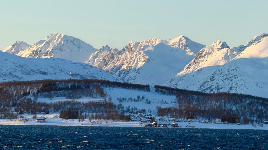 A small village on a snowy coast with blue water in the foreground and a large, sunlit mountain range behind.