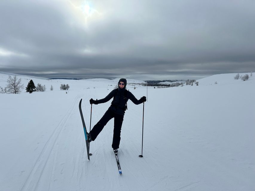 A person in black ski gear poses playfully on cross-country skis in a vast, snowy landscape under an overcast sky.