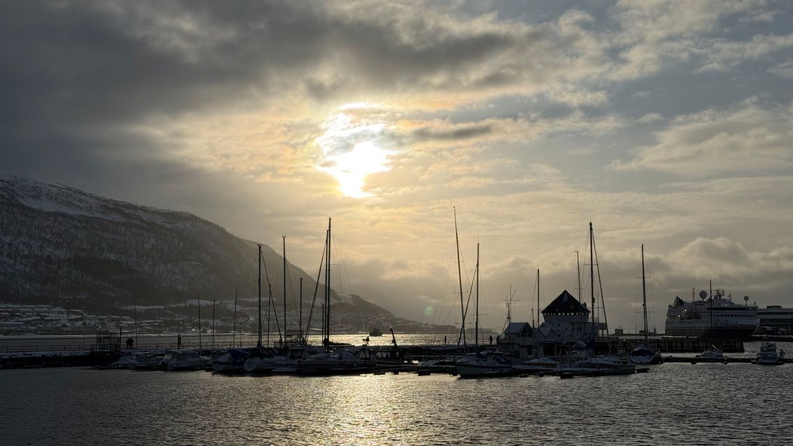 Sailboats docked in a snowy harbor at sunset, with light reflecting on the water and a mountain in the background.