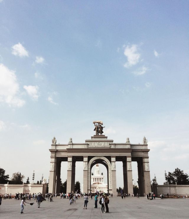 Inquadratura dal basso di un grande arco monumentale in una piazza affollata sotto un cielo luminoso con nuvole leggere.