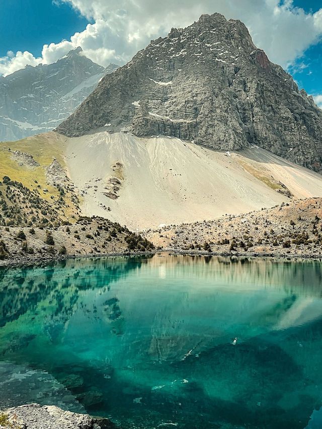 Un'imponente montagna rocciosa e un cielo nuvoloso si riflettono nelle acque immobili e turchesi di un lago alpino.