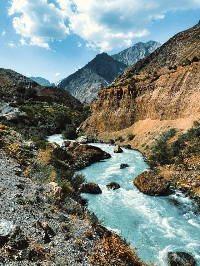 Un fiume azzurro scorre impetuoso attraverso un canyon roccioso di montagna sotto un cielo parzialmente nuvoloso.