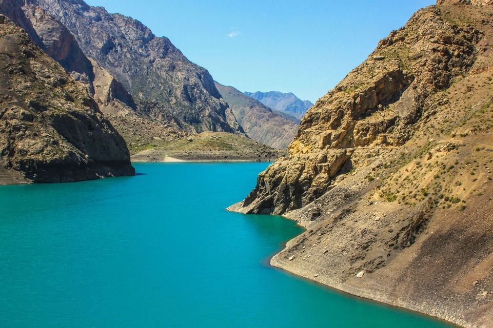 Un vivace lago turchese scorre attraverso un profondo canyon, circondato da ripide montagne rocciose sotto un cielo azzurro e limpido.