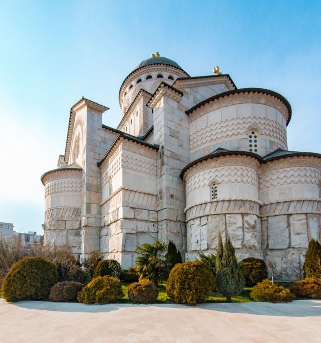 Una grande cattedrale di pietra bianca con una cupola centrale e intagli intricati, circondata da un giardino verde sotto un cielo azzurro e limpido.