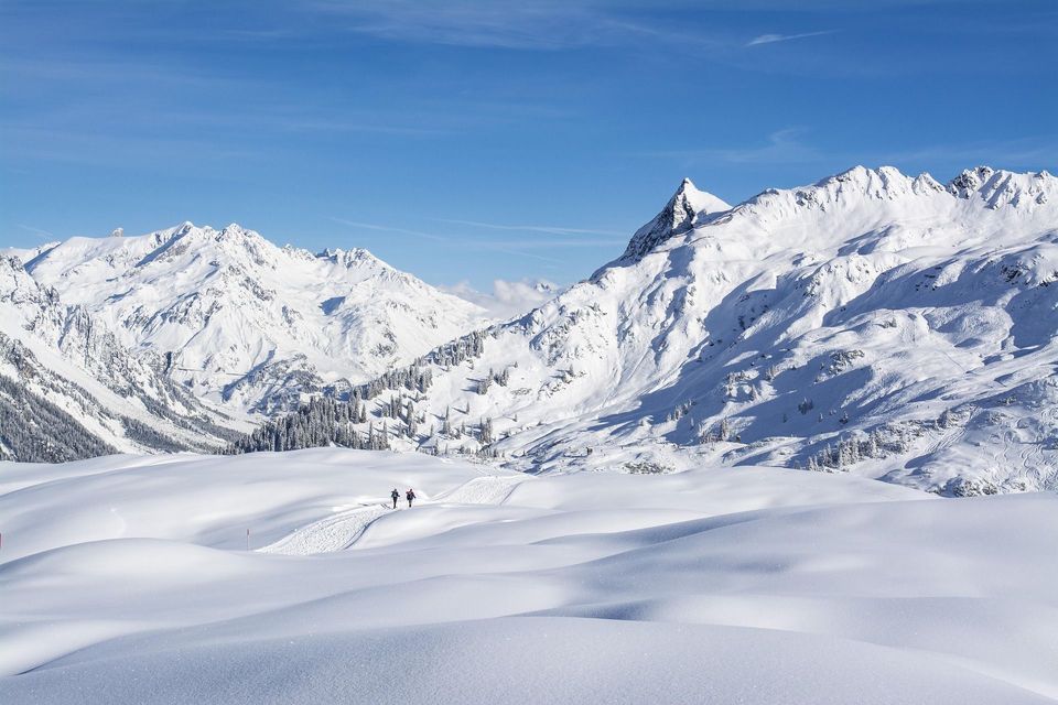 Un viaggio di gruppo WeRoad di due persone che camminano su un sentiero in un vasto paesaggio di montagne innevate sotto un cielo azzurro e limpido.