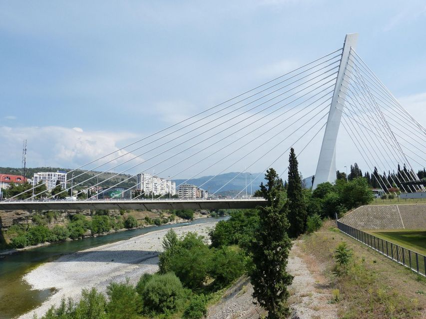 Un moderno ponte strallato bianco che attraversa un fiume poco profondo, con uno skyline cittadino e colline lontane sullo sfondo.