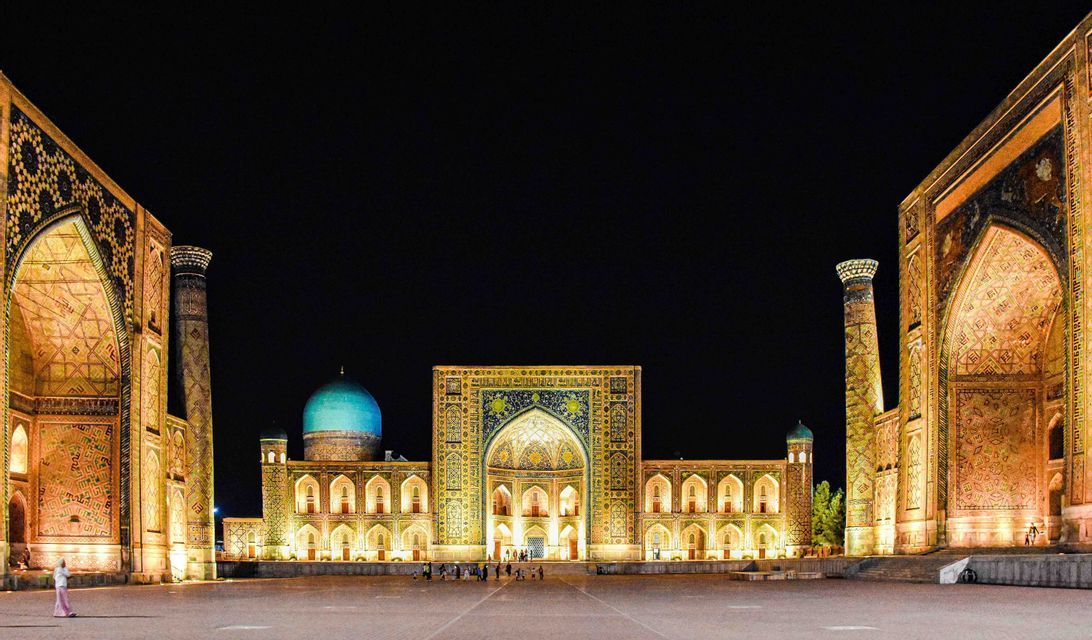 Un ornato complesso architettonico con cupola turchese e minareti è illuminato da luci dorate in un'ampia piazza di notte.