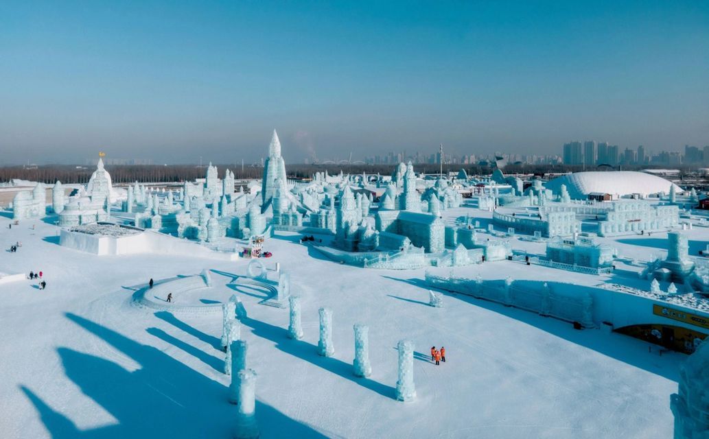 Vista aérea de un festival de invierno con grandes y complejas esculturas de hielo y nieve que cubren un paisaje nevado bajo un cielo azul despejado.