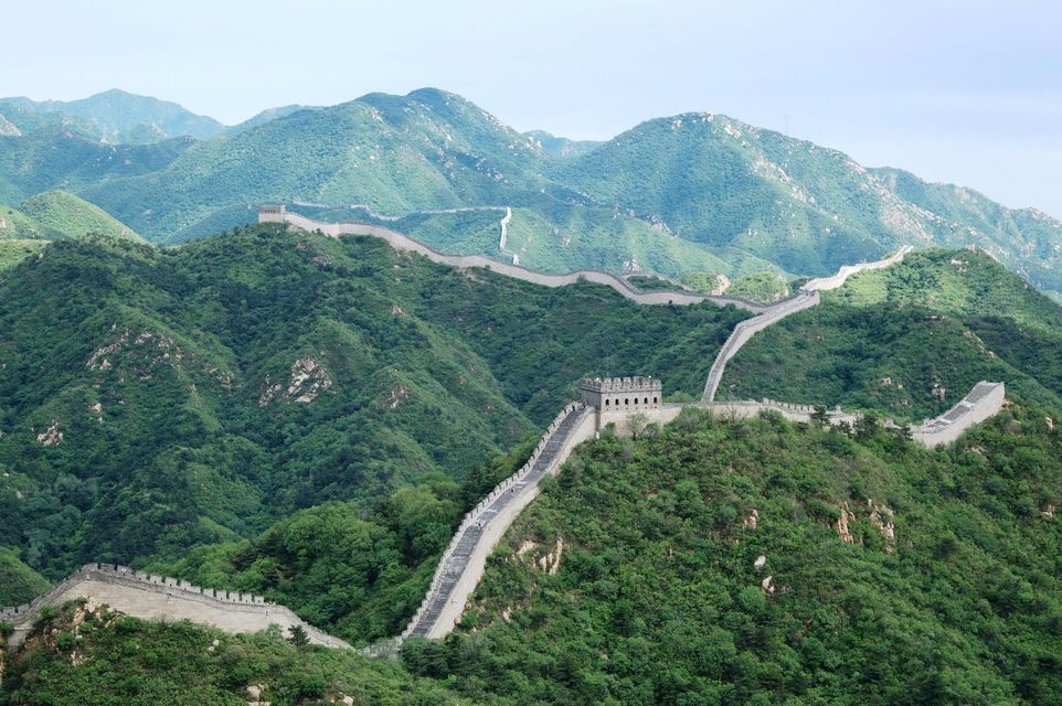 La Gran Muralla China serpentea a través de un vasto paisaje de montañas verdes y frondosas bajo un cielo pálido y nublado.