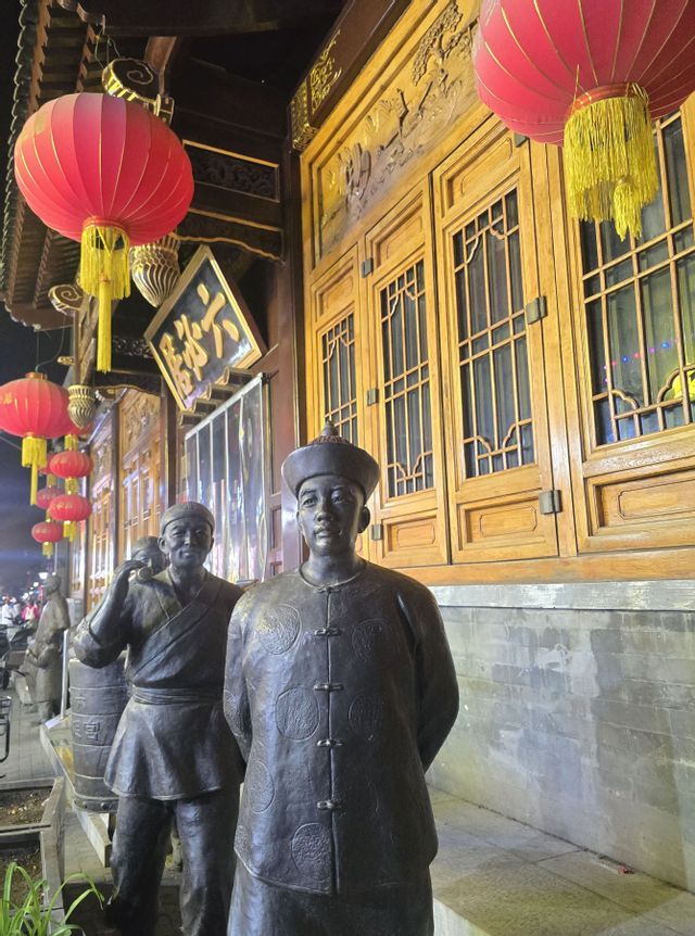 Dos estatuas de bronce de hombres con atuendo tradicional se encuentran fuera de un edificio decorado con farolillos rojos colgantes por la noche.